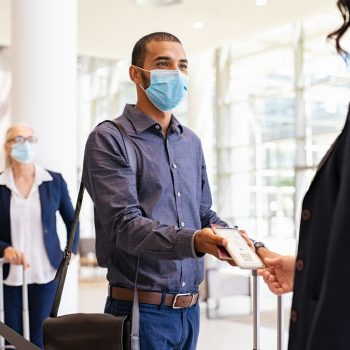 Indian passenger wearing surgical mask showing e-ticket to flight attendant at boarding gate. Young mixed race businessman showing boarding pass on mobile phone to air hostess while wearing protective face mask during covid pandemic. Multiethnic business man in a row with flight reservation hand the phoone to stewardess at airport keeping social distance.